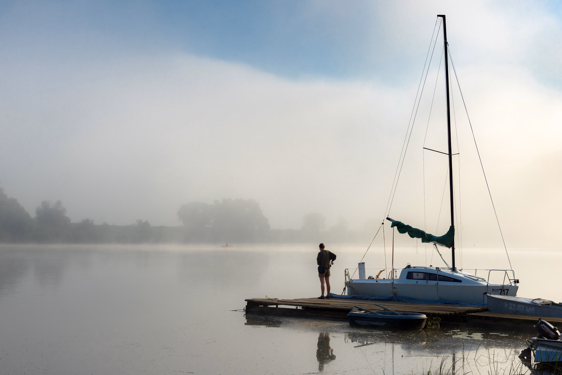 A person on a jetty by a sailboat in morning fog on the Oka River